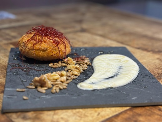 SIDRERÍA VIP. Nombre: Cachopito VIP. Un delicioso bocado que fusiona tradición y creatividad. El protagonista es un jugoso cachopo de tomate relleno de rulo de queso de cabra y cecina de León, logrando un equilibrio perfecto entre cremosidad y el sabor ahumado de la cecina. Descansa sobre una base de salsa roquefort enriquecida con cerveza Mahou, cuya textura y profundidad de sabor complementan cada capa del pincho. Un acabado con un toque de miel y frutos secos aporta un contraste dulce y crujiente, haciendo de este pincho una explosión de matices en el paladar. Tapa Mahou. ( PUEDE AMPLIARSE)